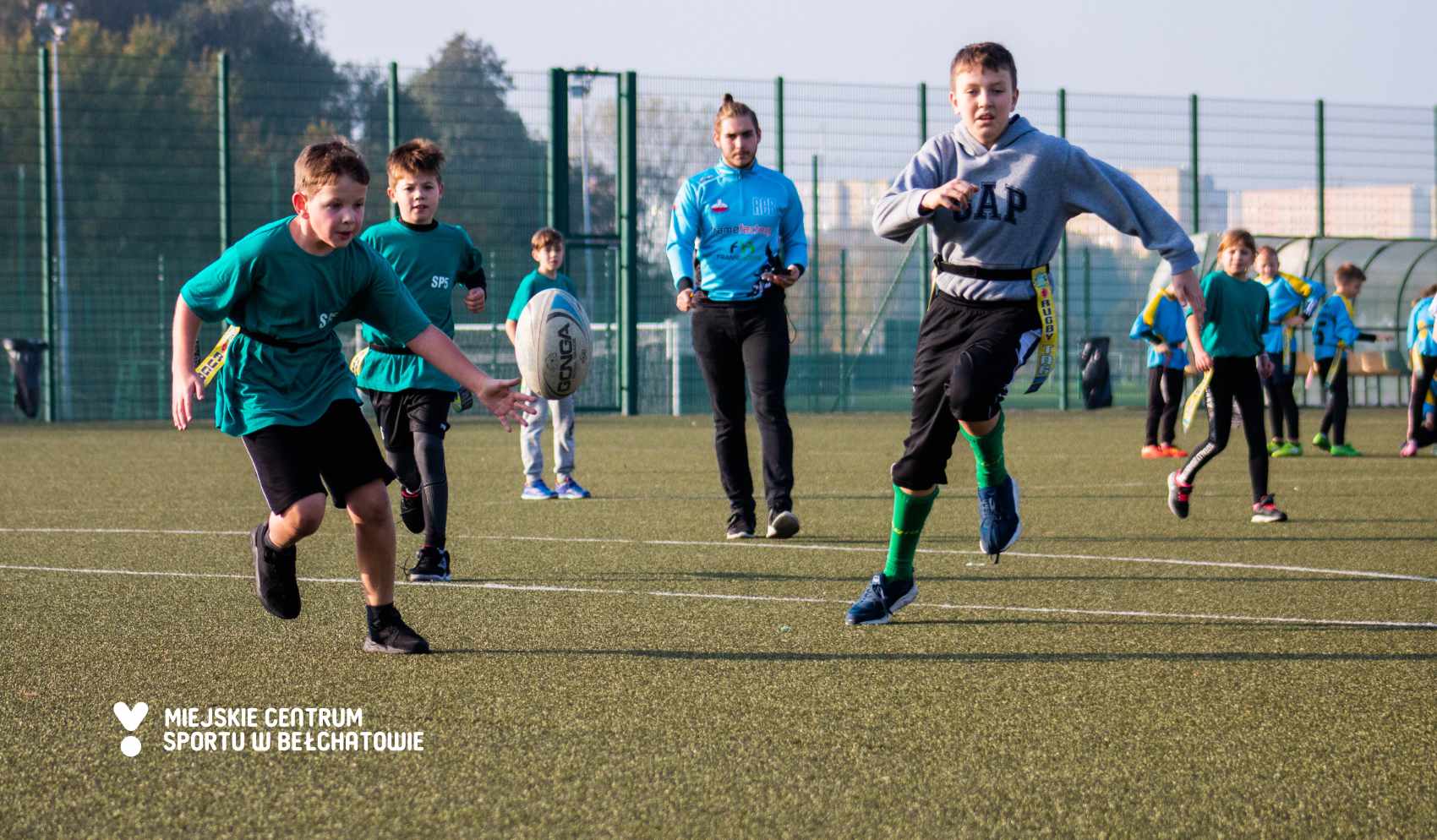 na zdjęciu widocznych dwóch uczestników turnieju Ogólnopolskiej Szkolnej Ligi Rugby Tag biegnących w kierunku piłki do rugby, a w tle widoczny sędzia i pozostali zawodnicy