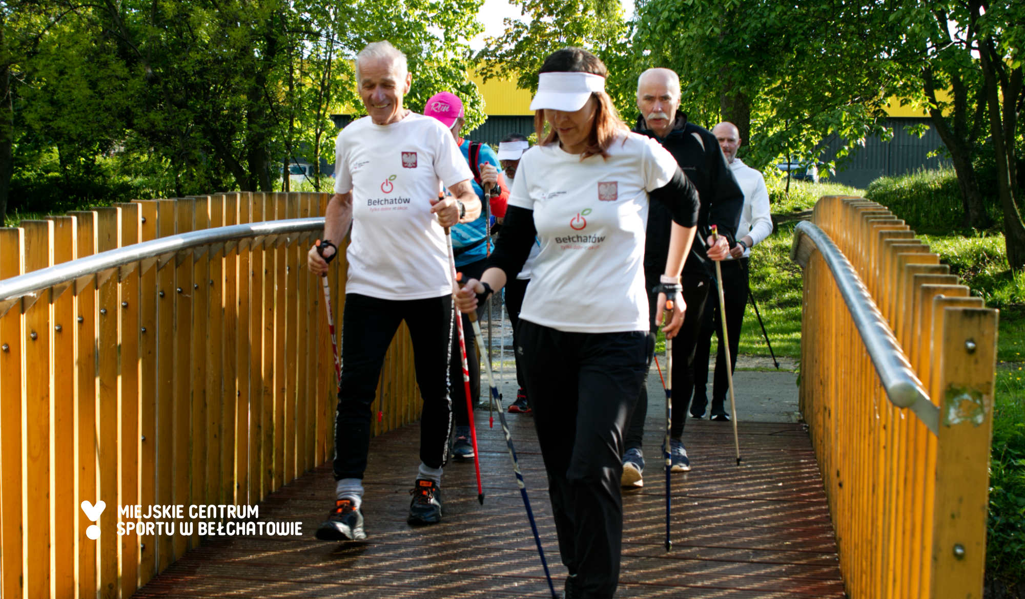 Na zdjęciu widoczni uczestnicy warsztatów Nordic Walking Bełchatów 2021 pod nazwą "Chodź po zdrowie!", którzy uczestniczyli w marszu rekreacyjnym idący przez kładkę nad rzeką w Parku Olszewskich